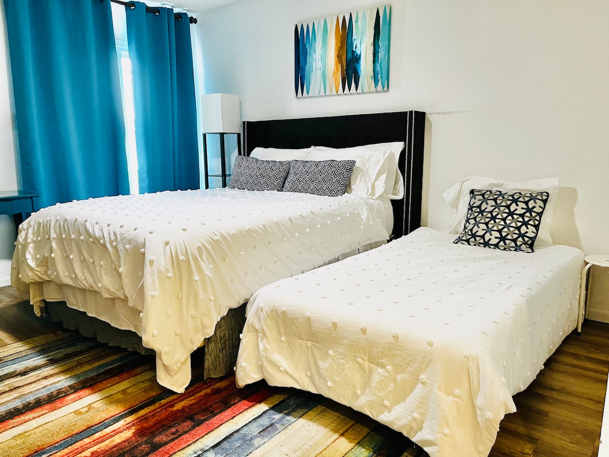 A spacious room is shown with two beds, featuring a larger bed and a twin-sized bed. Light-colored bedding contrasts with a colorful area rug. Tall curtains frame a window, allowing natural light to fill the space, and a piece of artwork hangs above the beds.