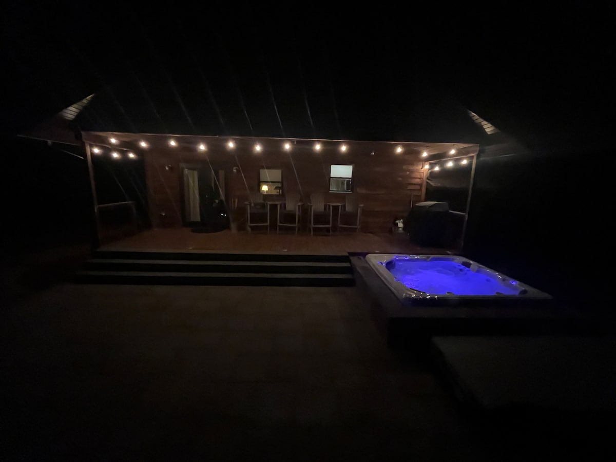 A covered back deck is featured, illuminated by soft string lights. A hot tub emits a gentle blue glow, while a dining set is arranged on the patio. Steps lead down from the deck, providing access to the outdoor space.