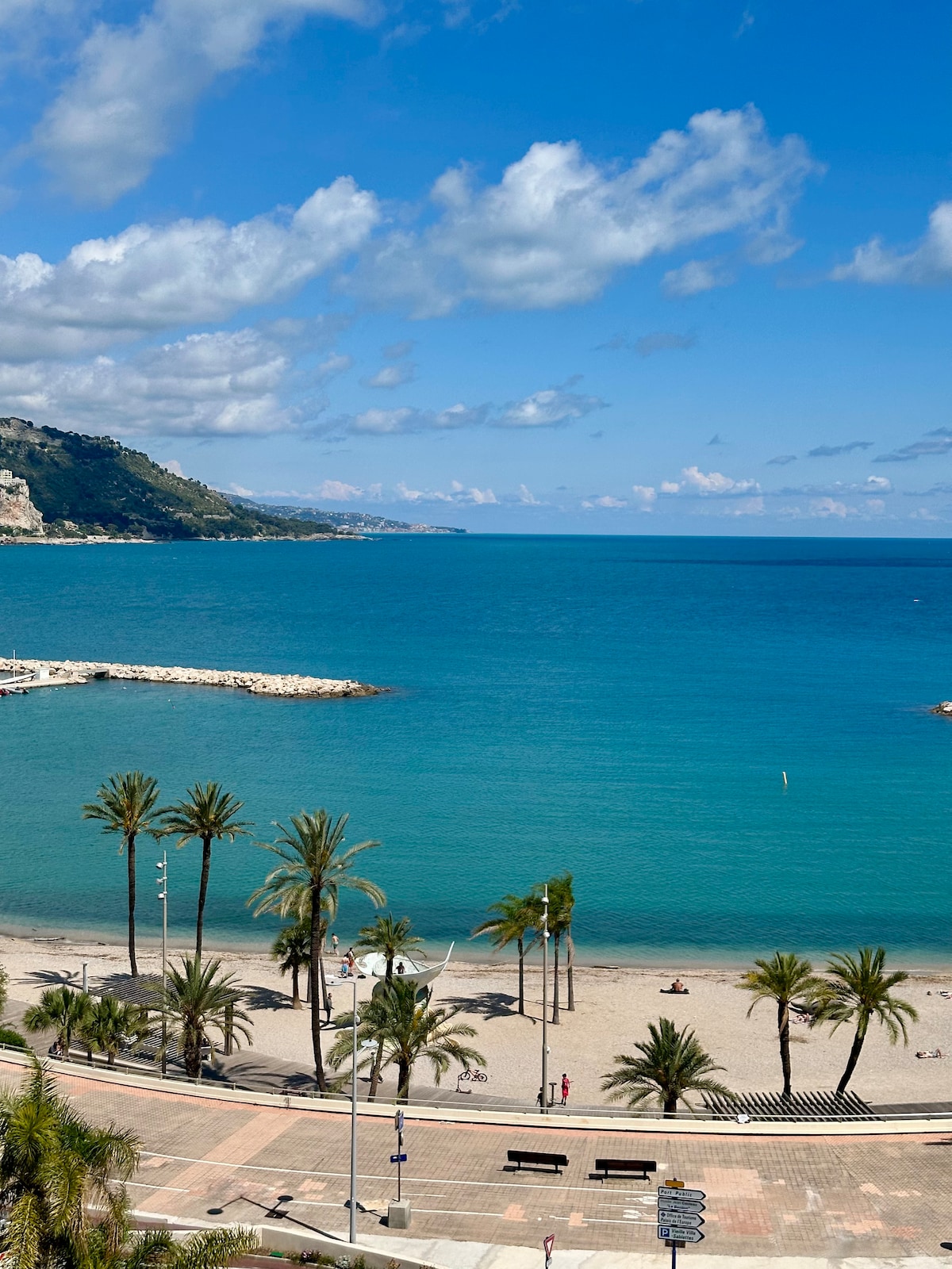 A serene beach scene is captured, featuring calm turquoise waters under a partly cloudy sky. Palm trees line the sandy shore, while a few individuals can be seen enjoying the open space. The coastline stretches into the distant hills, enhancing the coastal view.