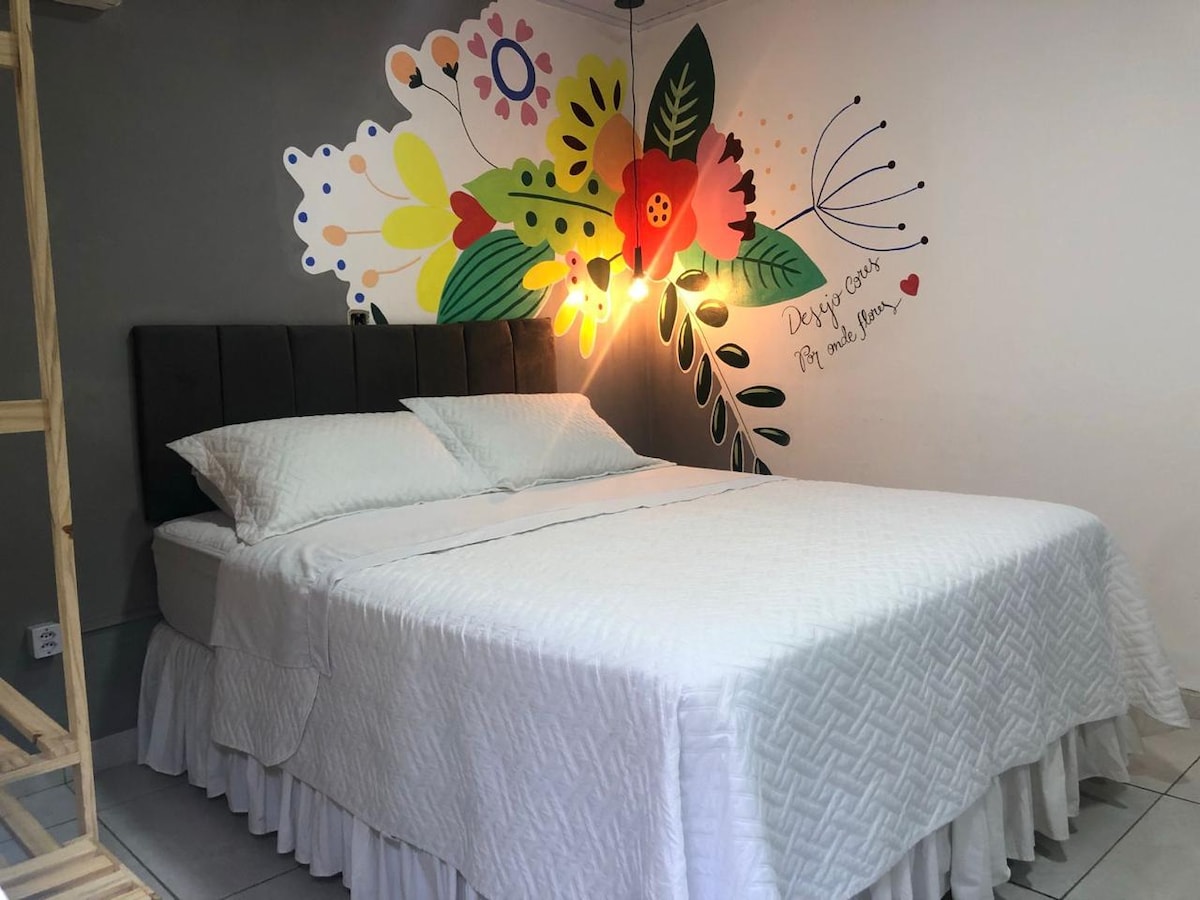 A spacious bed is covered with a white quilt. Colorful floral artwork is displayed on the wall above the bed, accompanied by a soft glow from an overhead light fixture. The room features simple, neutral tones and a tidy appearance.