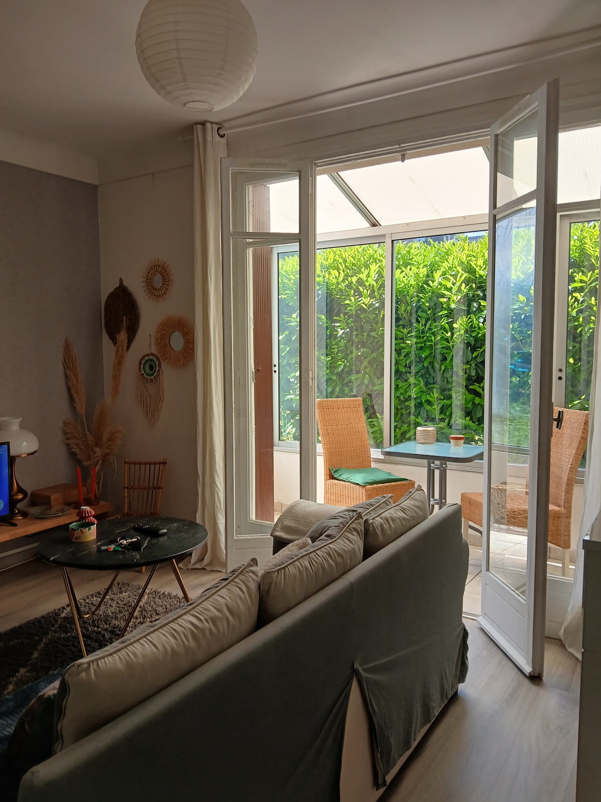 A comfortable living area features a soft sofa facing a glass door that opens to a light-filled loggia. The loggia is equipped with a small table and chairs, surrounded by lush greenery, providing a serene outdoor space.