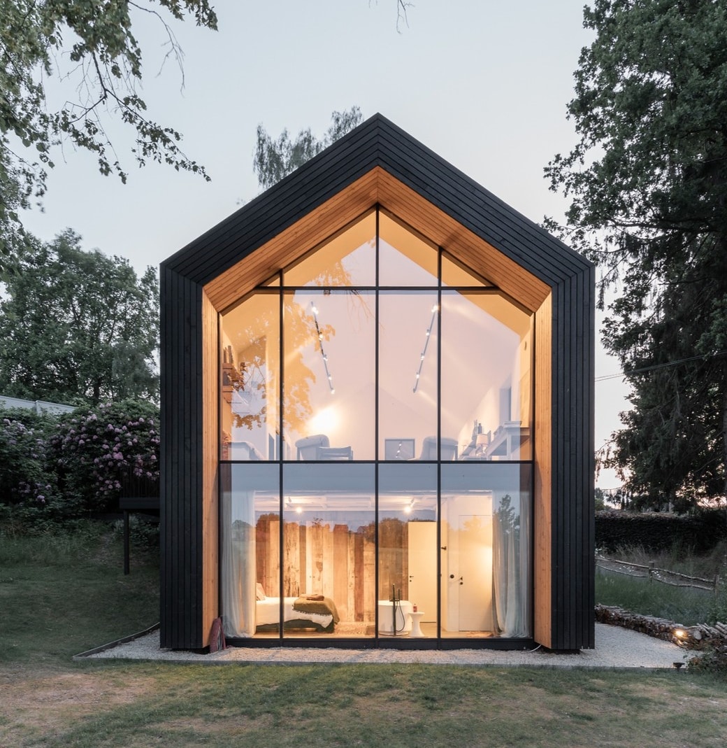 The modern cabin showcases a striking A-frame structure with large glass panels reflecting the soft evening light. Warm interior lighting illuminates the space, highlighting the natural wood elements and creating an inviting contrast with the dark exterior. Surrounding greenery adds to the serene atmosphere.