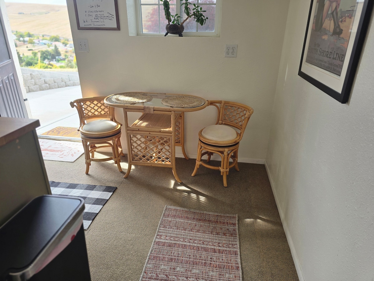 A small kitchenette area features a round rattan table with two matching chairs, offering a cozy spot for meals. Light enters through a nearby window, illuminating the simple decor and enhancing the inviting space.