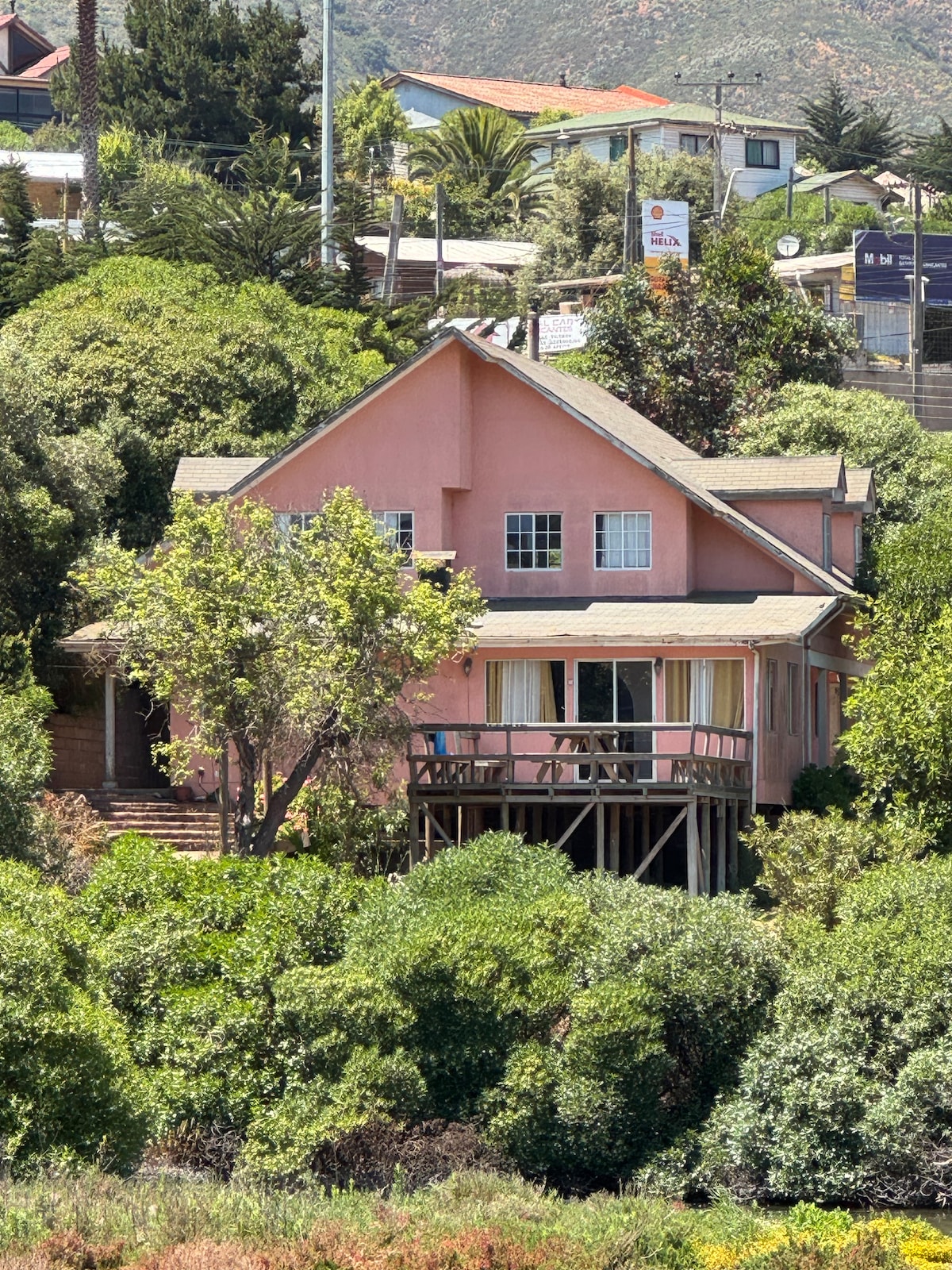 A charming pink house is nestled among lush greenery, featuring multiple windows that allow natural light to fill the interior. The elevated structure rests on stilts, enhancing the view of the surrounding landscape. A balcony can be seen, extending from the front of the house.