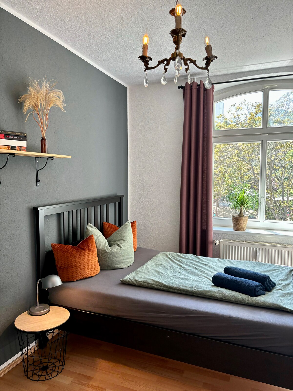 A cozy bedroom features a dark wooden bed adorned with decorative pillows in earthy tones. A green blanket is neatly spread across the bed, and a small bedside table holds a lamp. Natural light flows through the large window, which is dressed with deep purple curtains.