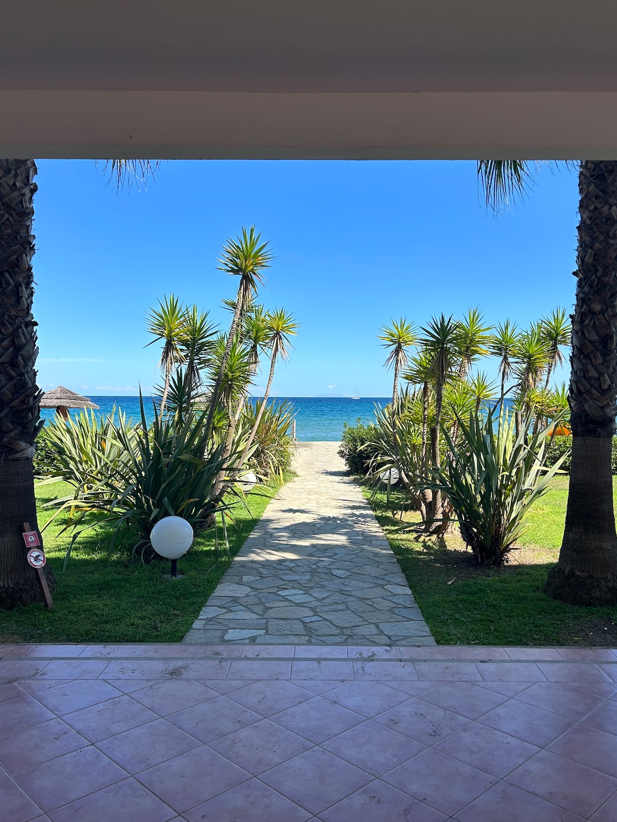 A path lined with greenery leads directly to the beach, framed by palms and other plants. The clear blue sea and sky are visible in the background, creating a serene coastal setting.