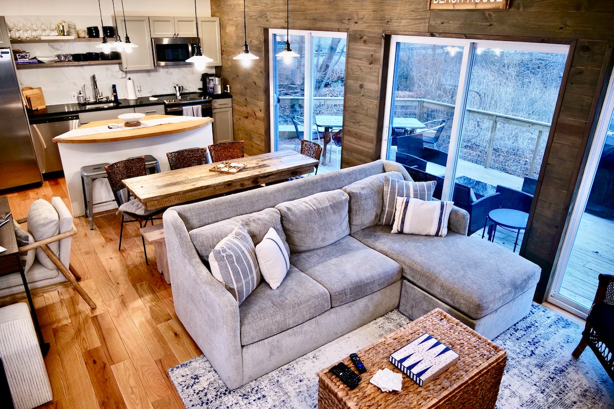 A spacious living area features a large sectional sofa adorned with decorative pillows, facing a wooden dining table. Natural light pours in through expansive glass doors, revealing a deck with outdoor seating. The kitchen is equipped with modern appliances and minimalistic cabinetry.