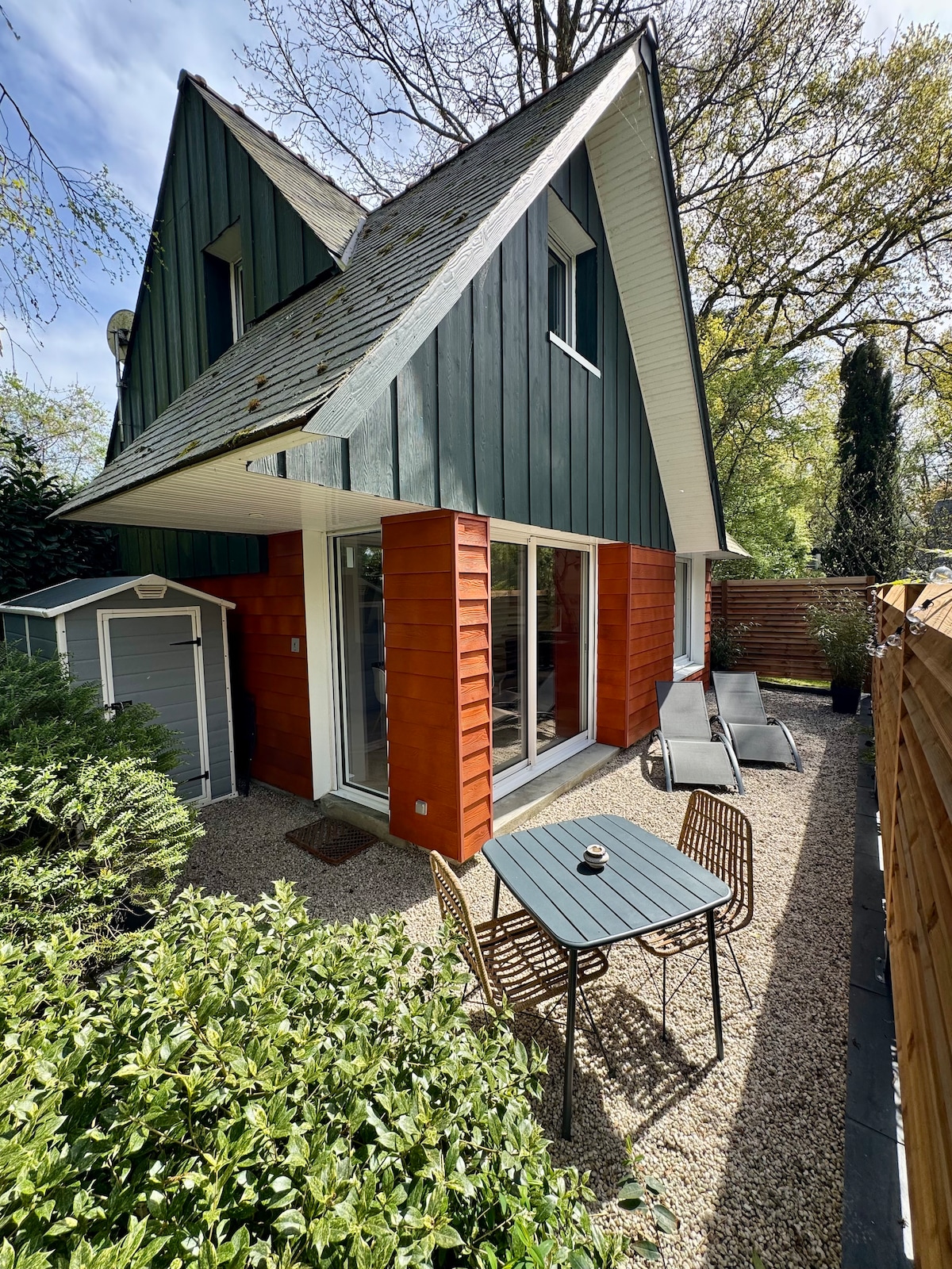 The exterior of the cottage features a blend of green and red paneling, surrounded by lush foliage. A gravel area includes two lounge chairs and a small table, providing a space for outdoor relaxation. A storage cabin is visible to the left.