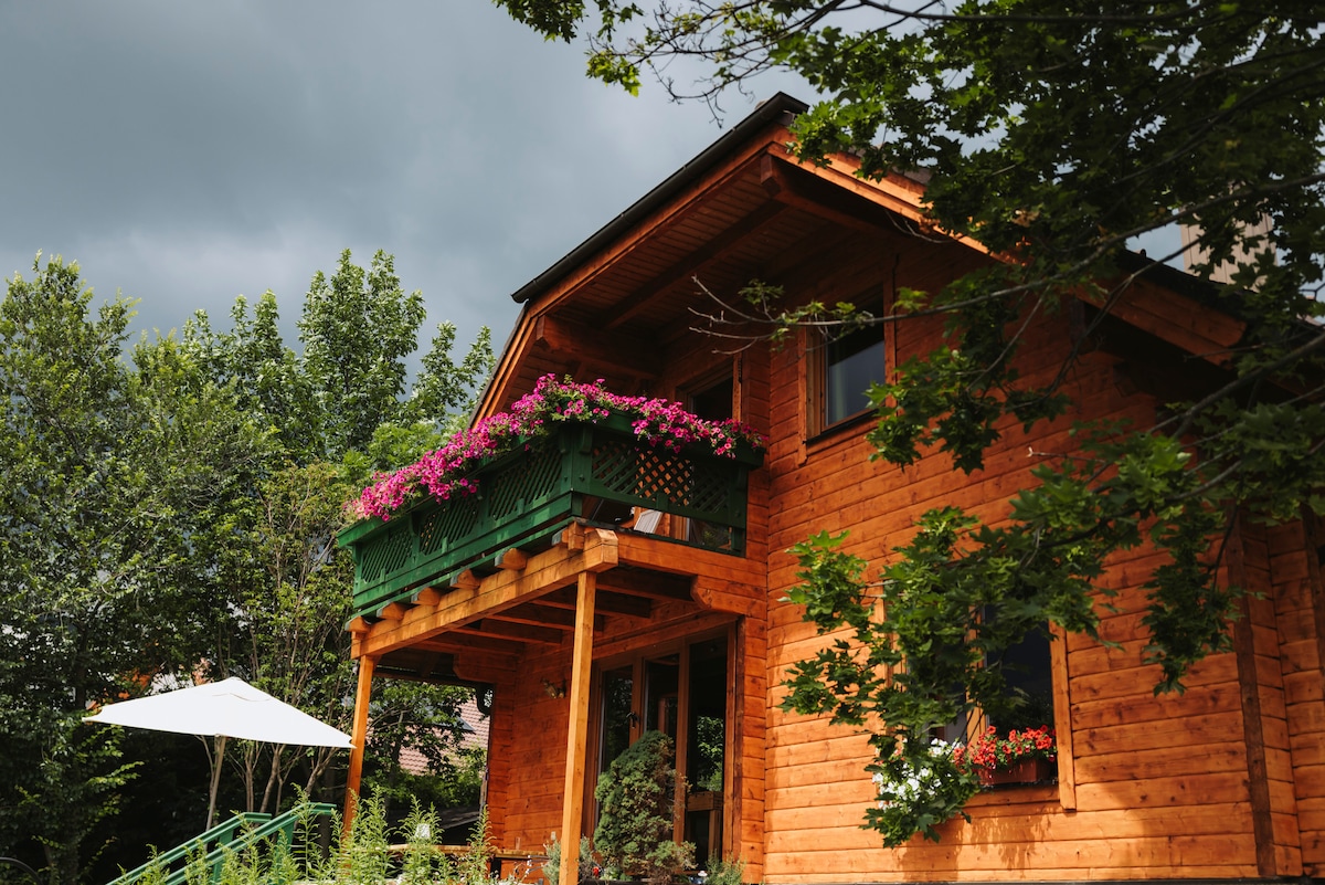 A wooden house features a balcony adorned with colorful flowers, surrounded by greenery. The entrance is framed by a canopy supported by sturdy wooden beams, while a white umbrella provides shade in the outdoor space below.