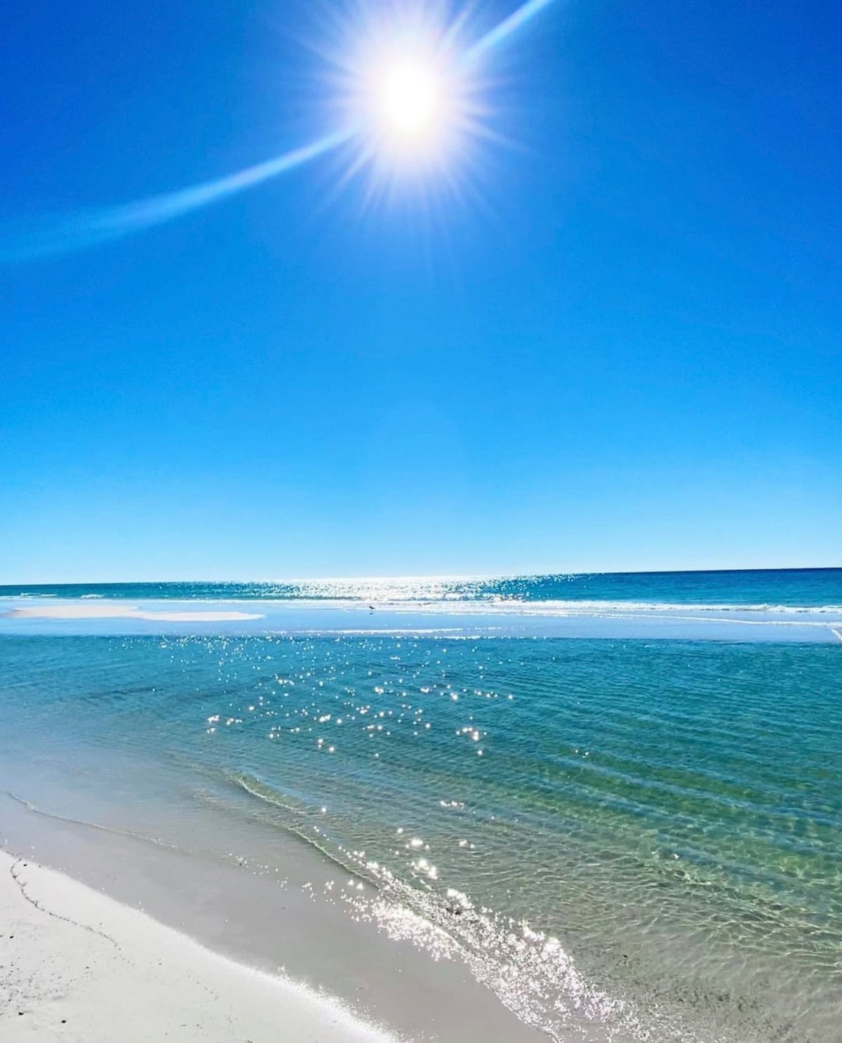 A serene beach scene is captured under a clear, blue sky, with the sun shining brightly. The water shimmers with reflections, and gentle waves lap at the soft, sandy shore. A calm atmosphere is evident, inviting peaceful moments by the sea.