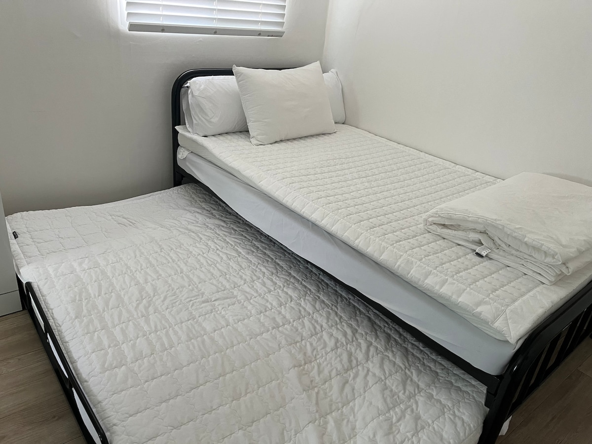 A minimalist sleeping area is presented with a spacious bed and a pull-out mattress positioned below. The bedding is neatly arranged in white, complemented by a folded blanket at the foot of the bed.