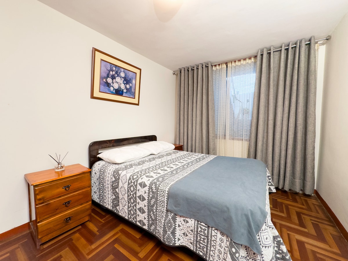 A cozy bedroom features a spacious bed dressed in patterned bedding, complemented by a bedside table and a wooden dresser. Natural light enters through a window adorned with sheer curtains, while gray curtains frame the window, creating a calm atmosphere.