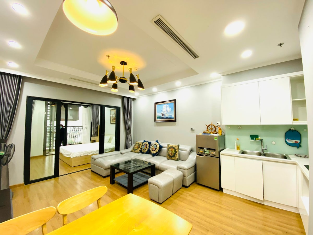 A modern living space is highlighted by a sectional sofa adorned with decorative pillows. A large glass door provides access to a balcony, while a dining table and fully equipped kitchen, featuring a refrigerator and sink, are visible. Soft lighting enhances the overall ambiance.