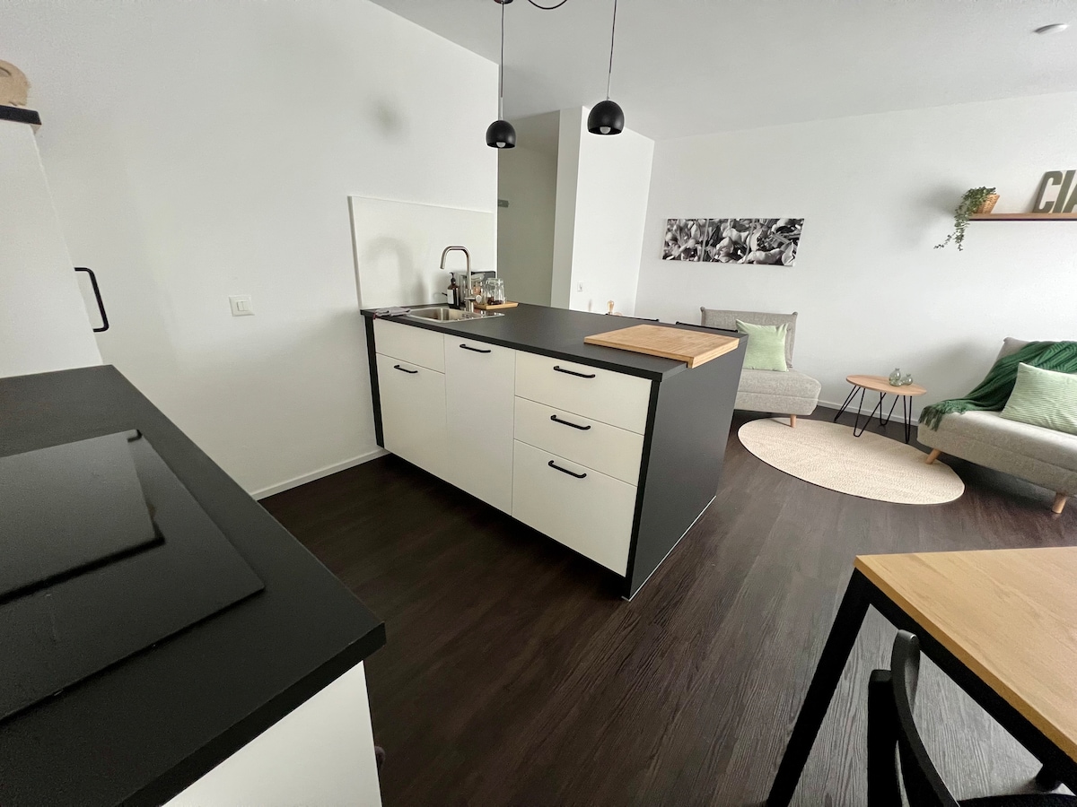 A modern kitchen is showcased, featuring a central island with a wooden chopping board. White cabinetry contrasts with dark countertops. In the background, a cozy seating area is visible with neutral-toned sofas and decorative accents, creating a functional and inviting space.