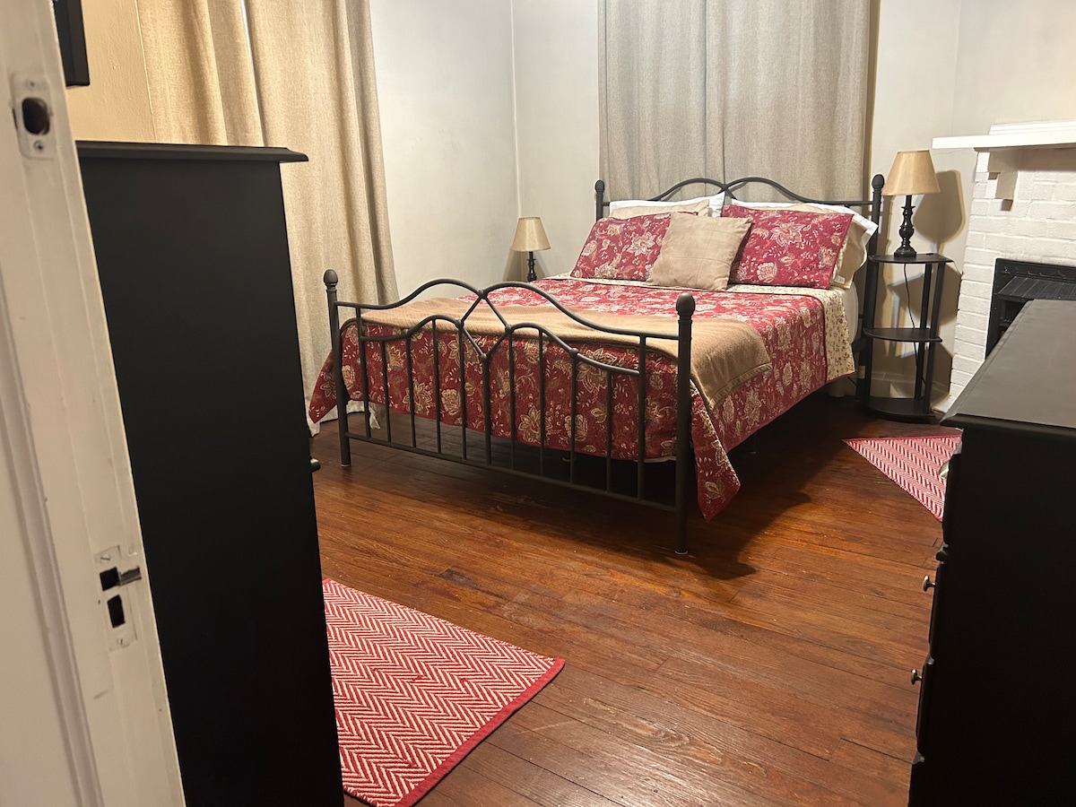 The Blackberry Room features a bed with an ornate metal frame, adorned with a floral quilt and decorative pillows. A nightstand with a lamp is positioned beside the bed, and a dresser stands against the wall. Natural light fills the room through the window covered with soft curtains, highlighting the warm wooden flooring.