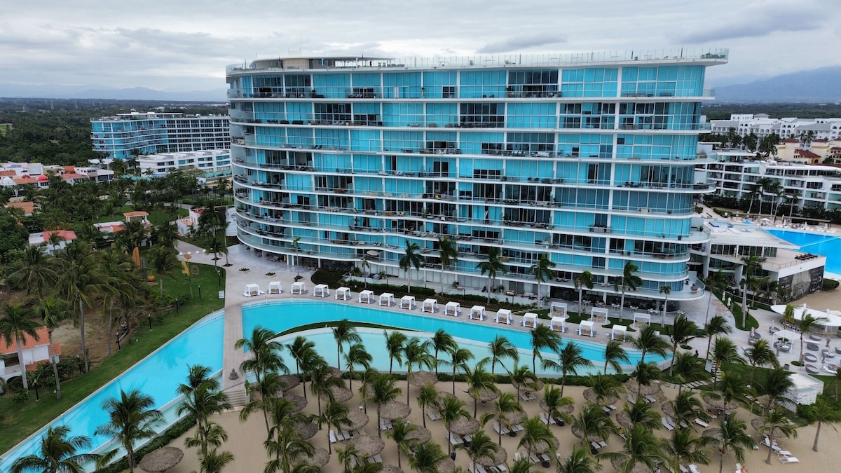 The resort features an impressive modern design with a curved, glass facade reflecting varying shades of blue. A large pool meanders through palm trees, surrounded by lounge chairs, leading to the expansive sandy beach area.