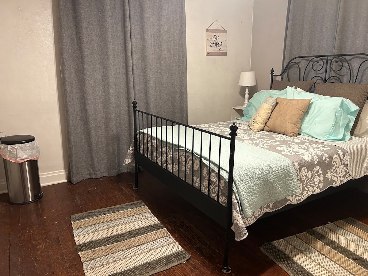 A tranquil bedroom features a metal bed frame with a light blue and gray comforter. The bed is adorned with decorative pillows. Light gray curtains frame a window, while a small lamp sits on a bedside table. A wastebasket and two area rugs complete the space.