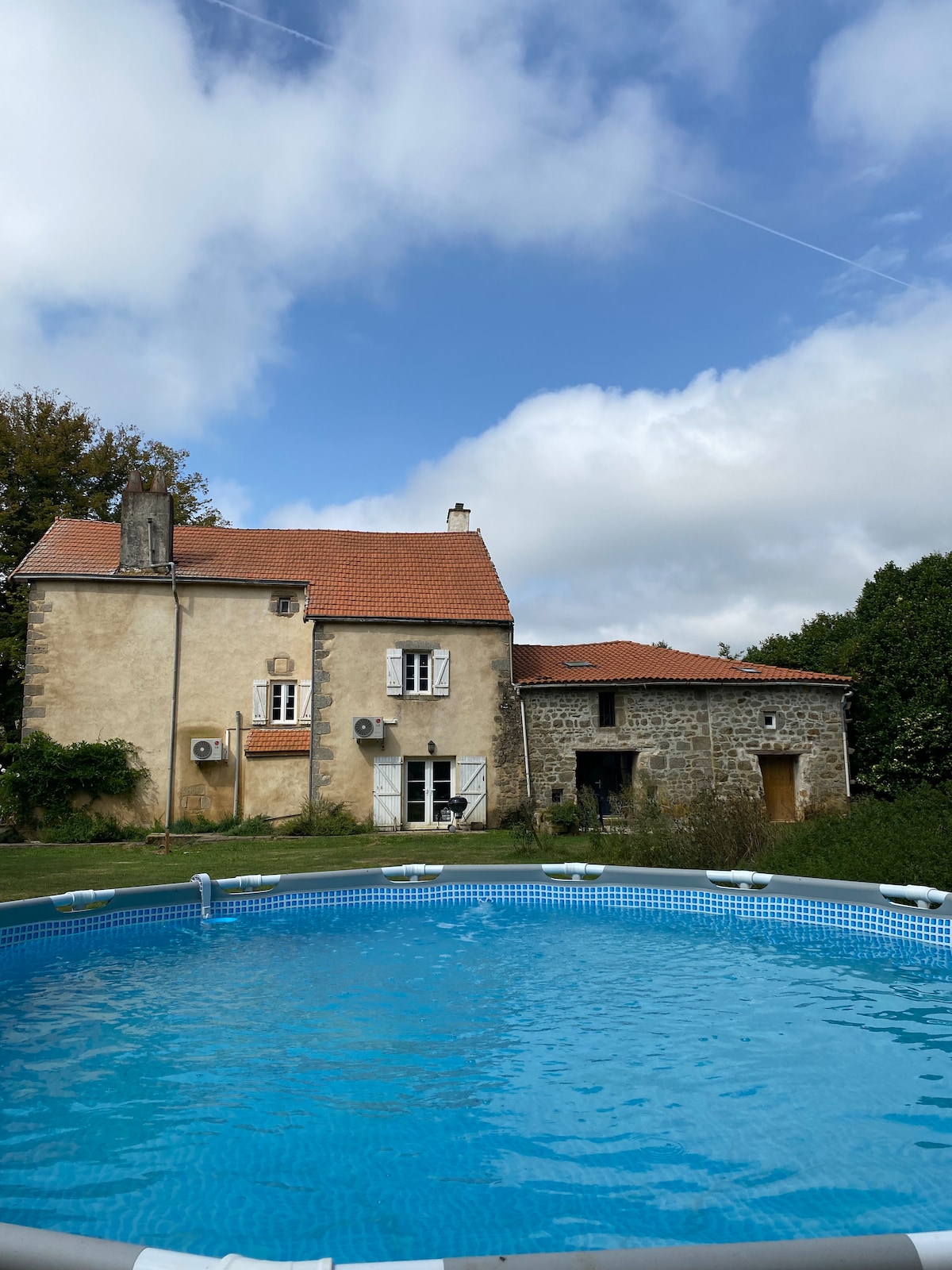 The cottage is depicted from an outdoor vantage point, showcasing its rustic exterior with a red-tiled roof. A circular above-ground pool is prominently featured in the foreground, reflecting the sky amidst scattered clouds, while the background includes lush greenery surrounding the property.