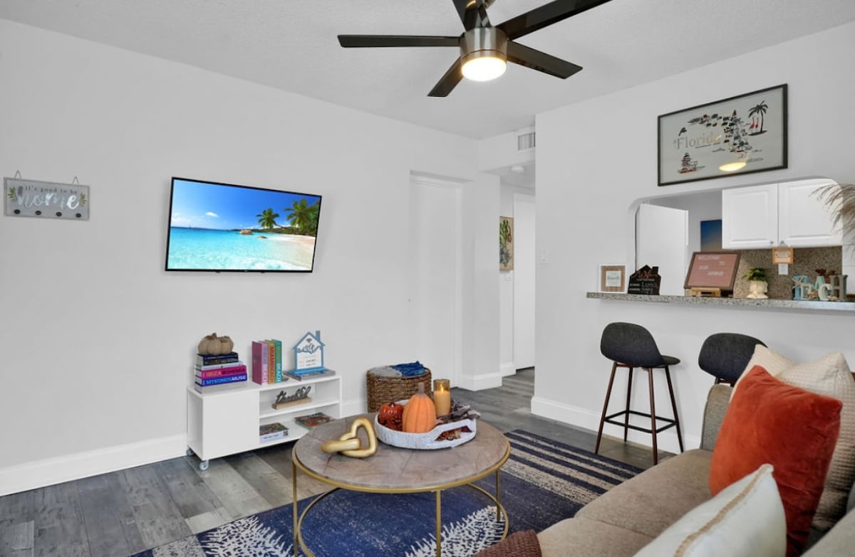 The living area features a soft sofa with colorful cushions and a round coffee table at its center, adorned with decorative items. A wall-mounted TV displays a serene beach scene, while a ceiling fan gently rotates overhead. An open kitchen can be seen in the background.