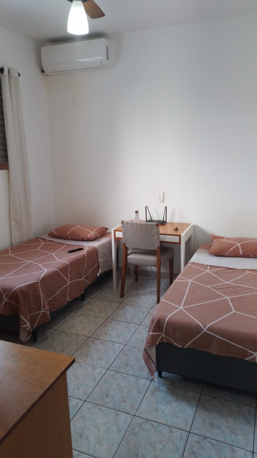 A tidy room features two twin beds with neutral, patterned bedding. A small desk with a chair occupies a corner, and a window allows natural light to illuminate the space. A ceiling fan and air conditioning unit are visible for climate control.