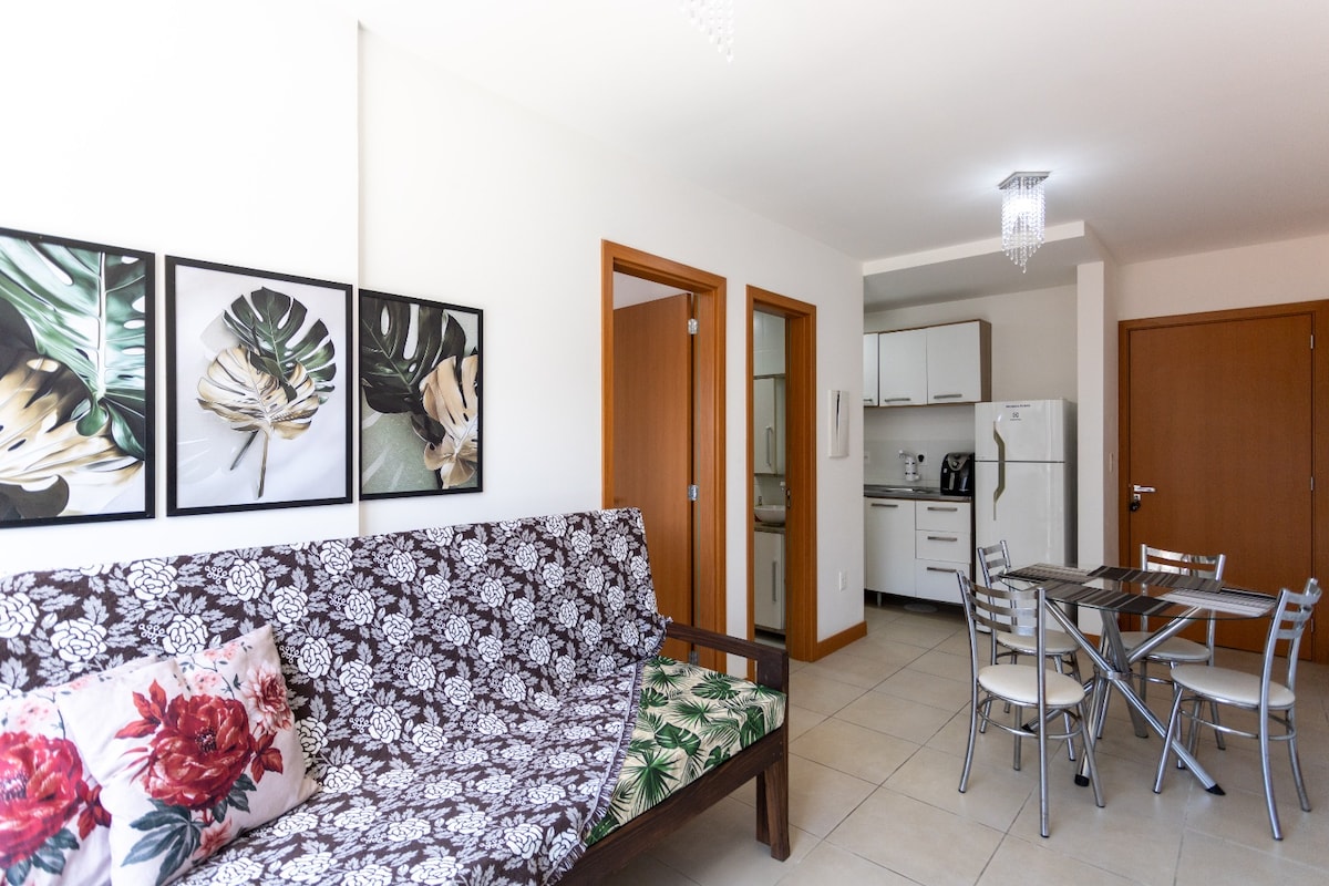 A modern living area features a comfortable sofa adorned with floral-patterned cushions. Adjacent to the seating area, a small dining table with metal chairs is visible. The kitchen area is equipped with essential appliances, while framed botanical art adds a touch of nature to the walls.