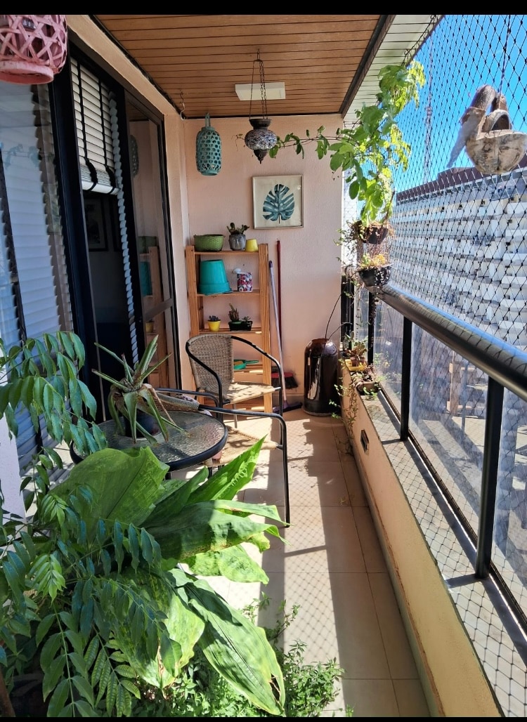 A narrow balcony is depicted, featuring potted plants and greenery along the edges. A small round table and two chairs are present, creating a cozy seating area. Decorative lanterns hang above, while a piece of wall art adds a touch of style, with ample natural light illuminating the space.