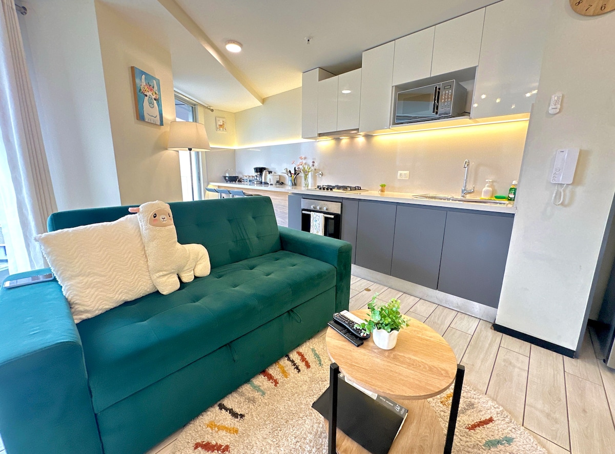 A modern kitchen is presented with sleek cabinetry and a stovetop. A cozy green sofa with a decorative pillow is positioned in the living area, accompanied by a small wooden coffee table. Natural light streams through windows, enhancing the inviting atmosphere.