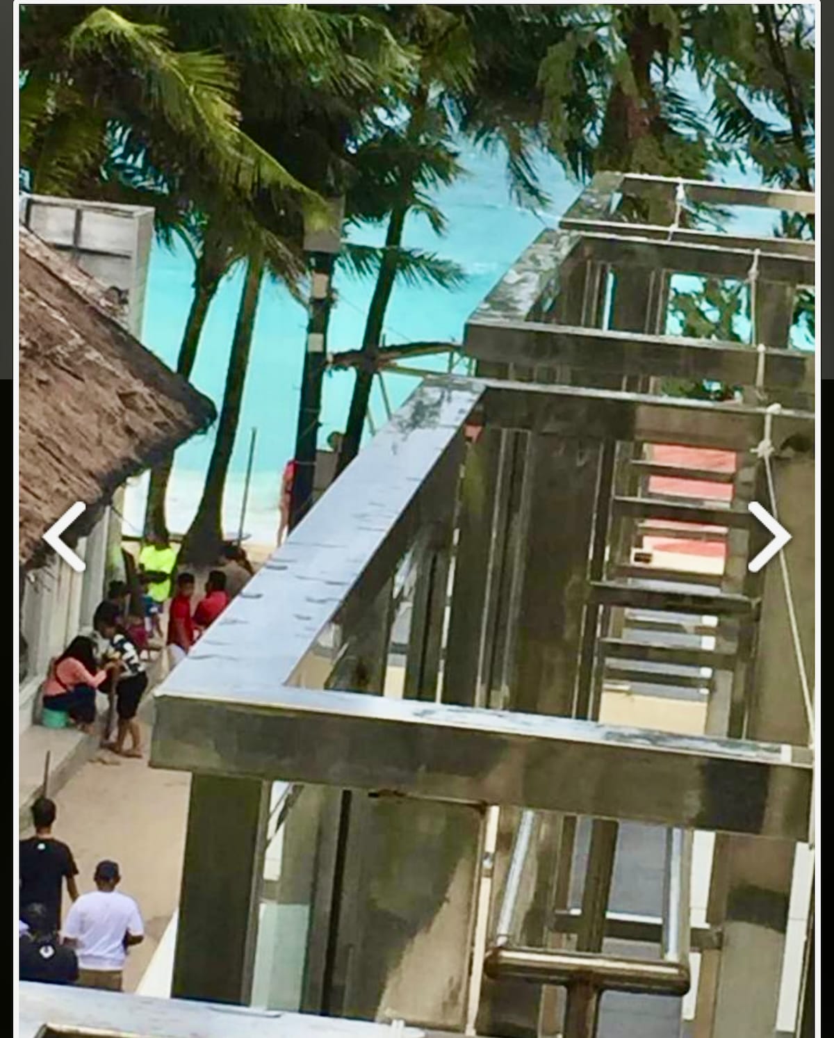 A view of a beachside pathway is captured, with tropical palm trees lining both sides. A glimpse of the turquoise water is visible in the background, while several people are seated along the walkway, enjoying the surroundings.