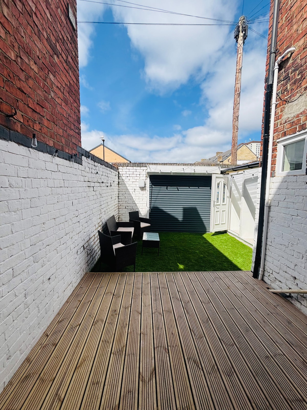 A private outdoor space features a wooden deck with seating arrangements. Green grass is visible, bordered by white brick walls and a storage unit. The sky above displays scattered clouds, providing ample natural light to the area.