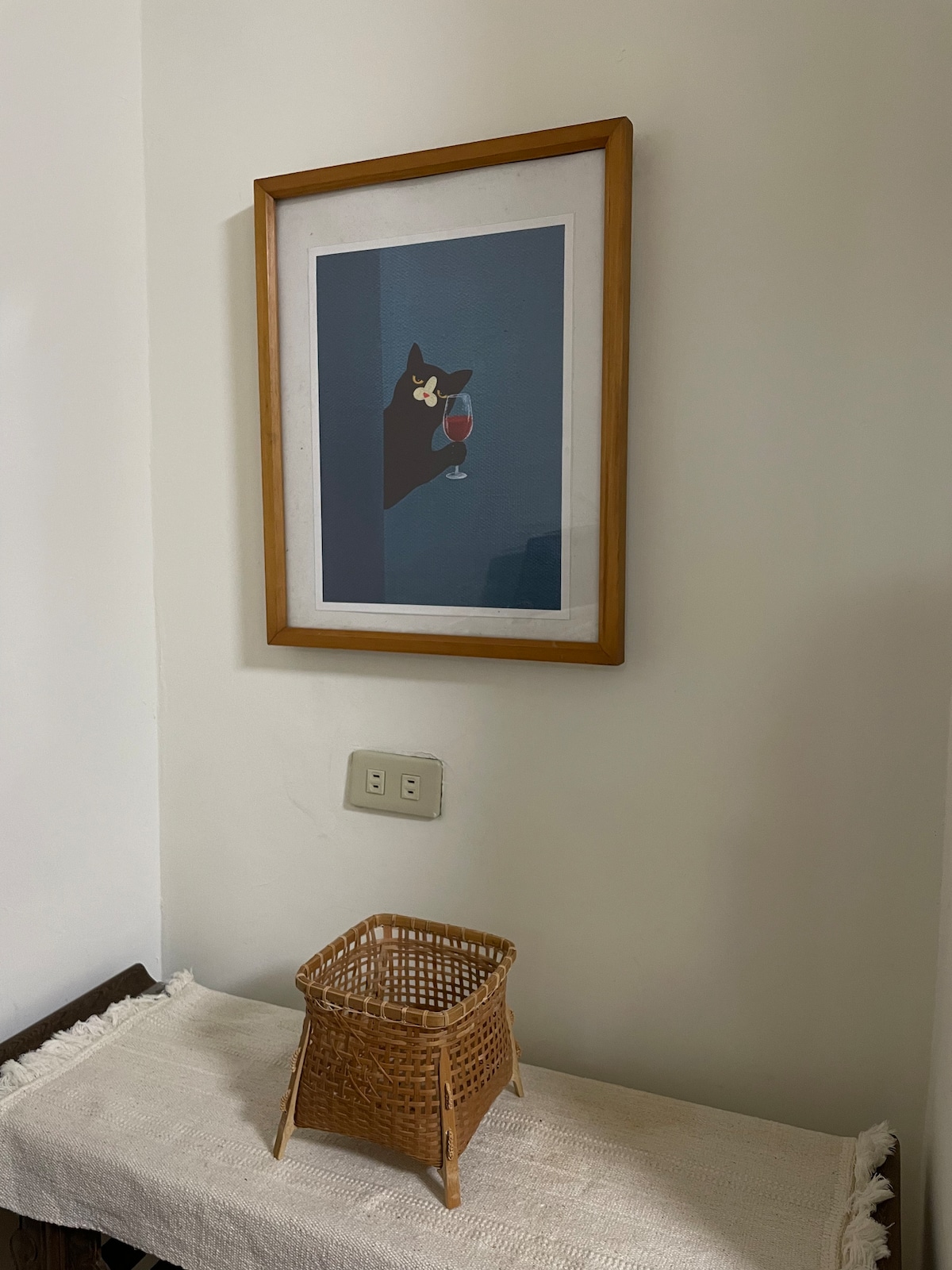 A framed artwork depicting a black cat holding a glass is mounted on a light-colored wall. Below, a woven basket sits atop a textured white cloth, adding an element of warmth to the space.