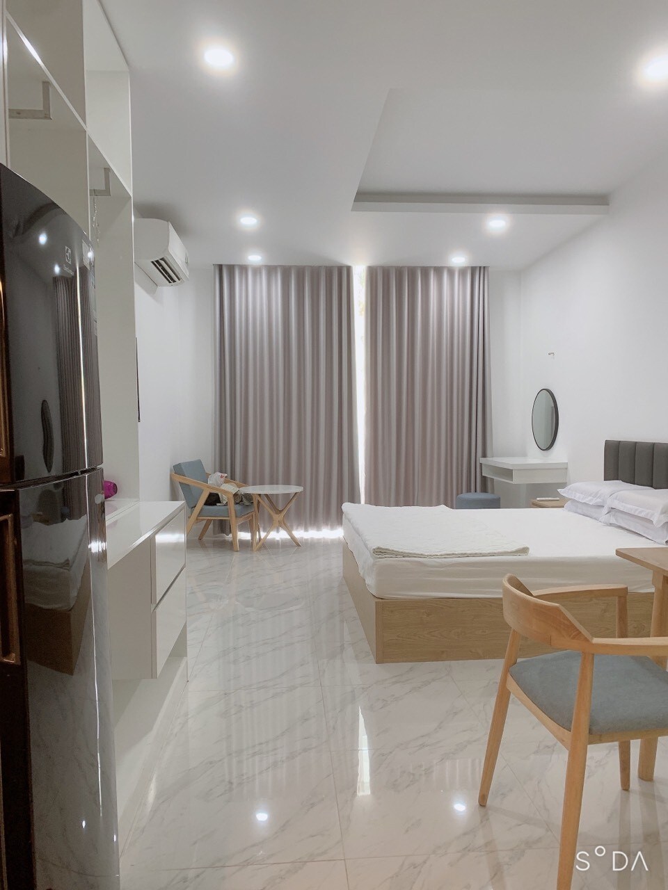 The studio features a spacious bed opposite a wooden dining table and chairs. Floor-to-ceiling curtains frame a window, allowing natural light to brighten the room. A compact kitchen area is visible along one wall, complemented by a sleek refrigerator and modern decor.