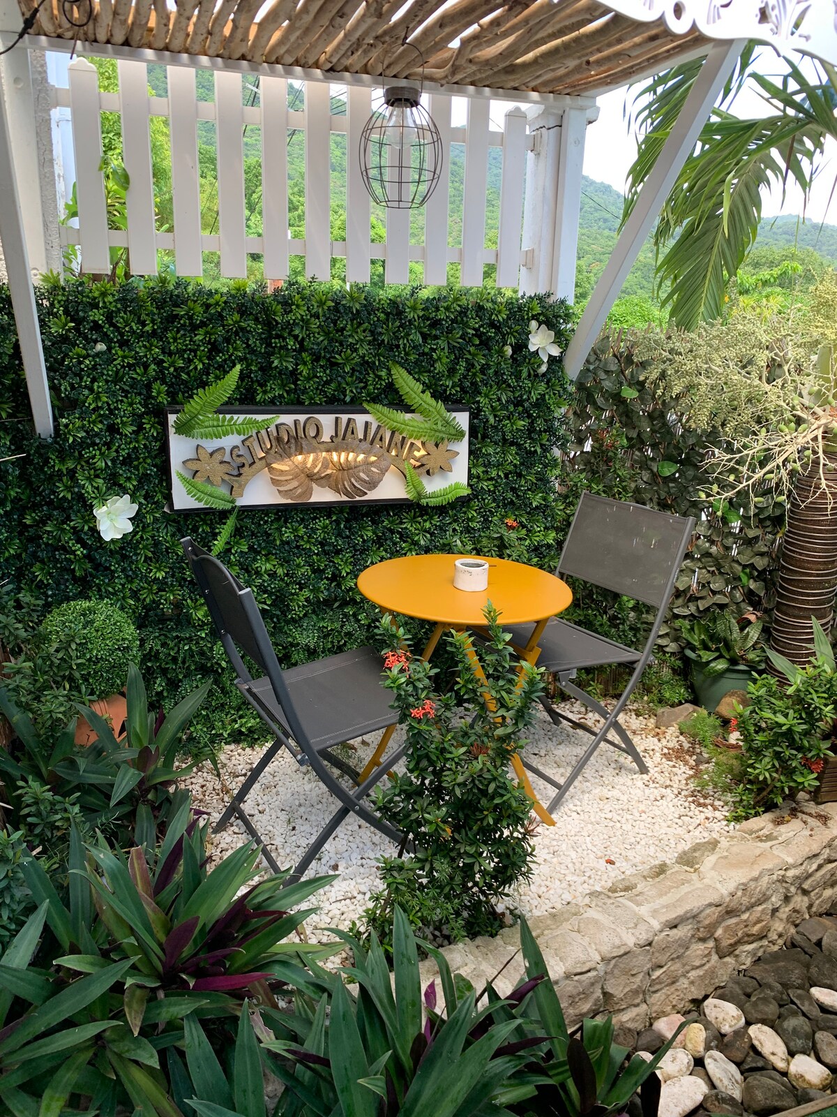 A small outdoor seating area features a round orange table accompanied by two gray chairs. The space is surrounded by lush greenery, including climbing plants and decorative elements, creating a tranquil spot for relaxation. A sign reading 'Studio Janais' is affixed to a green wall.