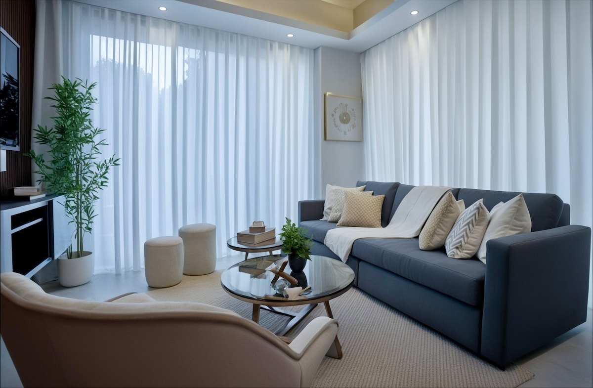 A modern living area features a dark sofa adorned with assorted cushions, positioned beside a stylish round coffee table. Soft white curtains allow natural light to filter through, while a potted plant adds a touch of greenery. A neutral area rug complements the decor.
