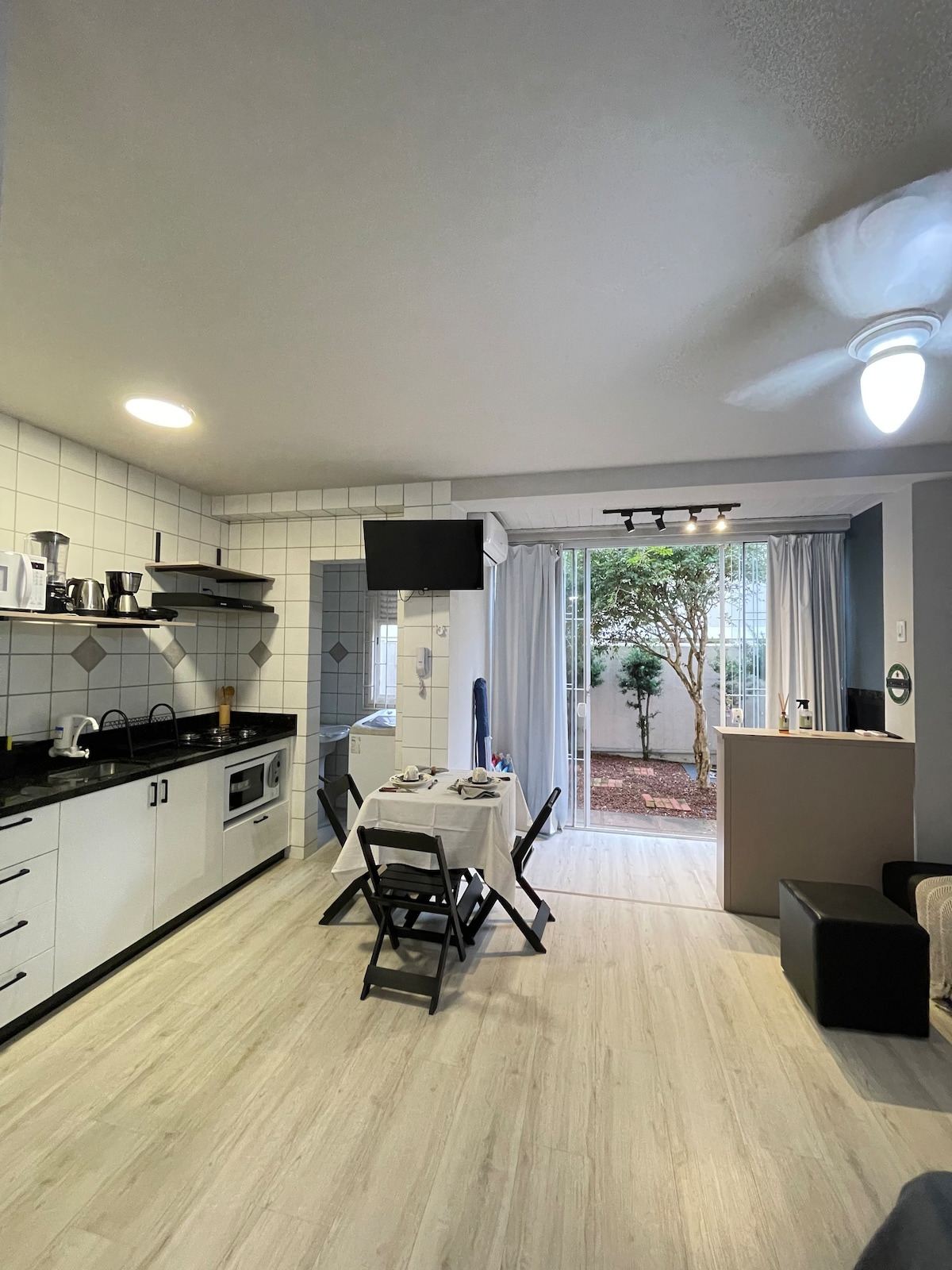 A bright kitchen area features modern appliances, including a stove and microwave. A dining table with two folding chairs is positioned nearby. Large glass doors open to a garden space, allowing natural light to fill the room. A ceiling fan is visible, providing air circulation.