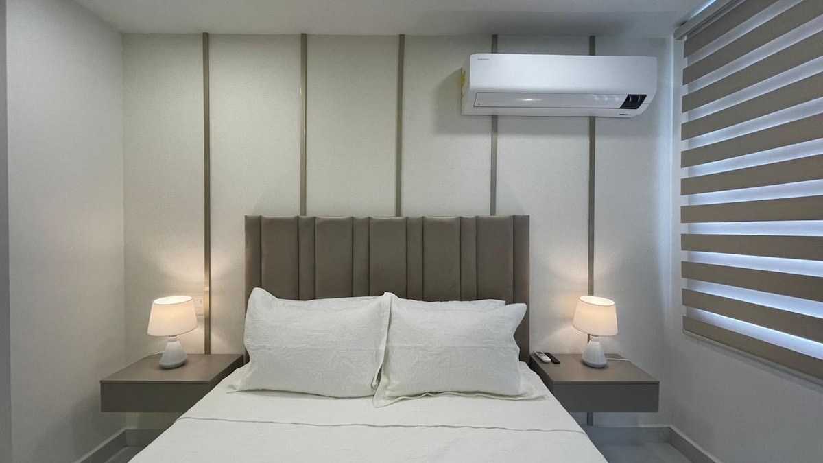 A spacious sleeping area features a modern upholstered bed with two soft pillows. Nightstands with lamps sit on either side, providing warm lighting. A window with light-filtering shades allows natural light to enter, while an air conditioning unit ensures a comfortable environment.
