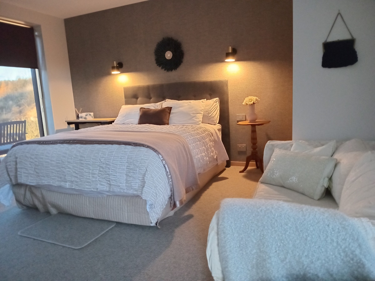 A spacious guest room features a plush bed with a light blanket and decorative pillows. A small side table with a vase, wall sconces, and a cozy seating area are included, while natural light filters through a window with a dark blind.
