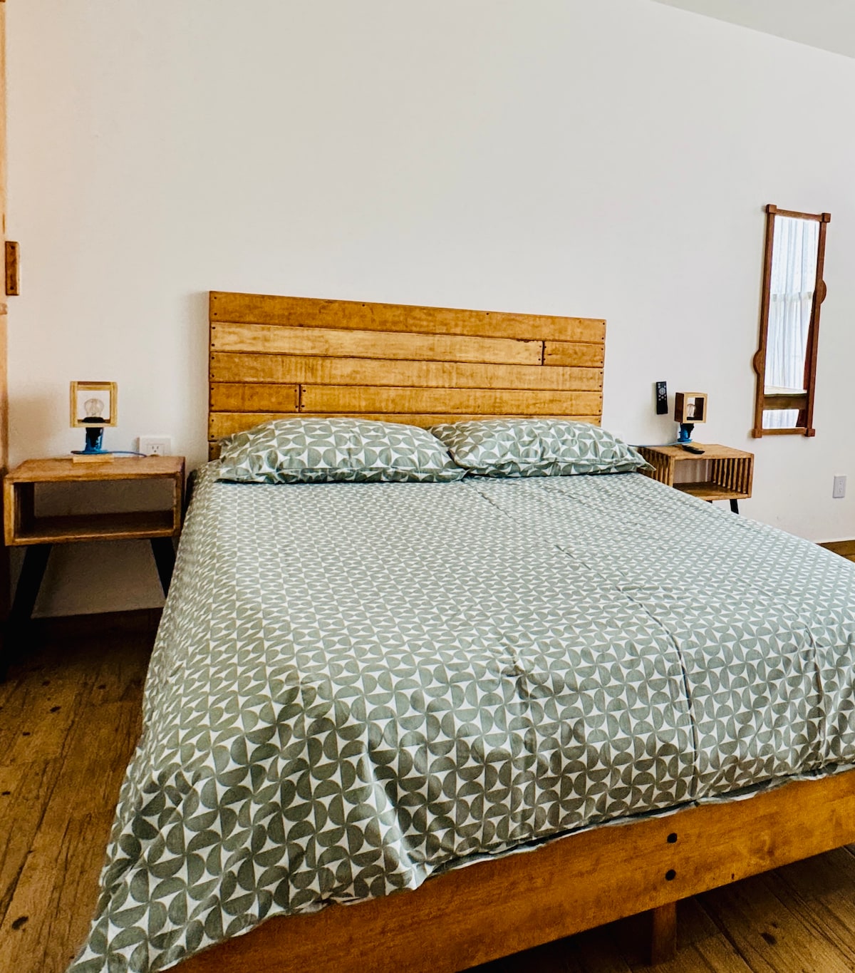 A spacious bed is centered in the room, dressed in a geometric patterned duvet. Two bedside tables, each featuring a lamp, are positioned on either side. A natural wood accent wall and a large mirror enhance the simplicity of the decor.