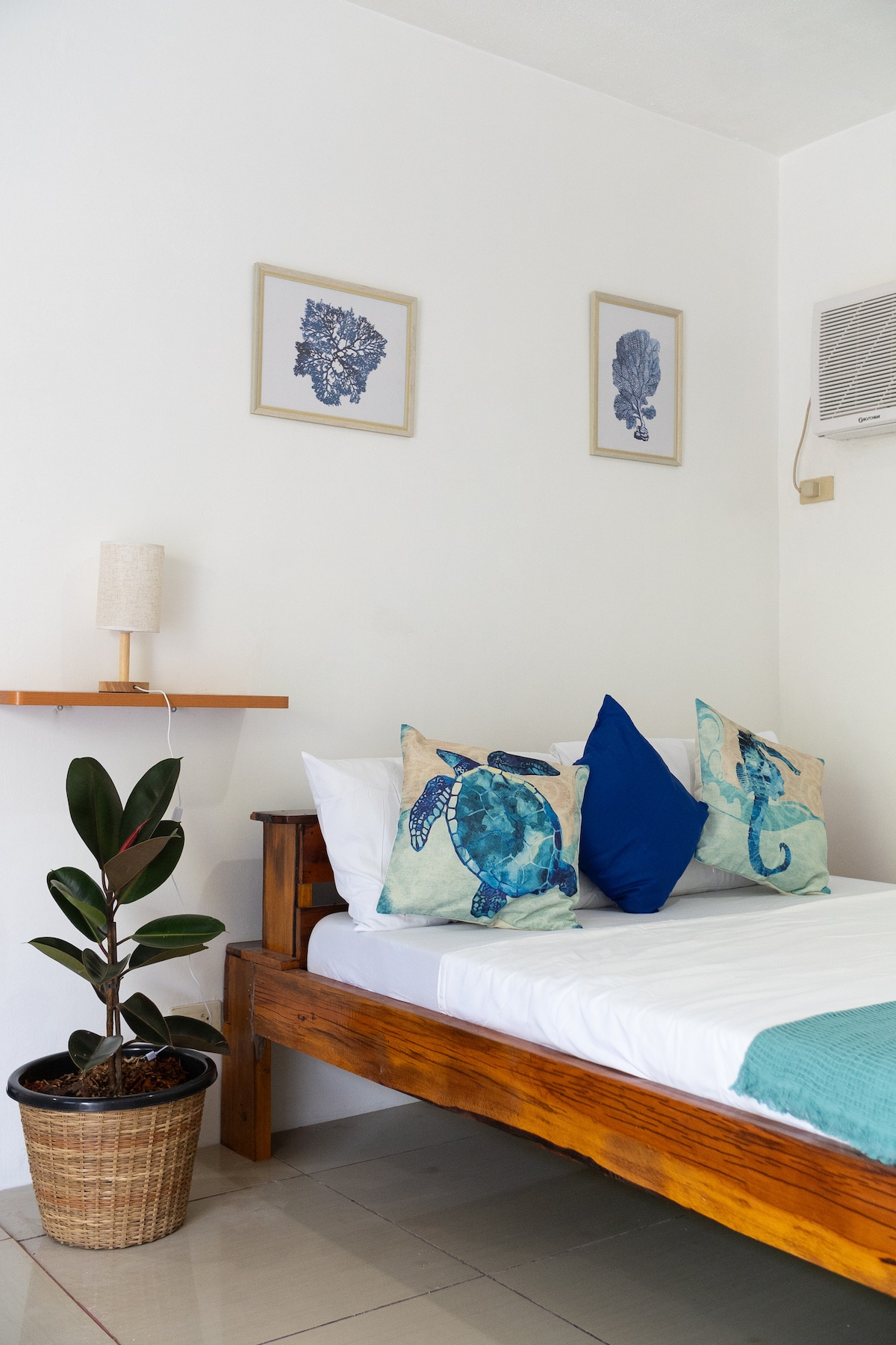 A cozy sleeping area features a spacious bed adorned with two decorative pillows showcasing marine-themed designs. The wall displays three framed artworks of coral, while a small decorative plant sits in a woven basket, adding a touch of nature to the room.