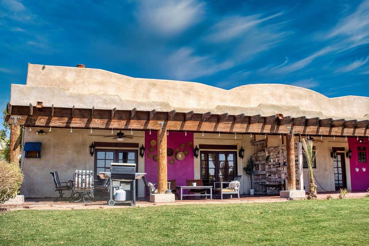 A spacious outdoor patio features a rustic wooden roof supported by beams. Comfortable seating is arranged alongside a grill, while bright pink walls and charming decor enhance the outdoor setting. The surrounding grassy area adds to the tranquil atmosphere of the property.