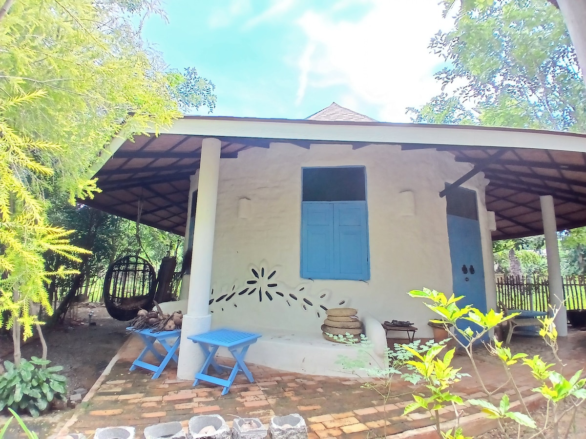 The exterior of the cob cottage is embraced by a spacious covered porch, featuring wooden support beams and a thatched roof. Light blue window shutters and a decorative element on the wall add charm. Blue chairs are placed on the brick patio, surrounded by lush greenery.