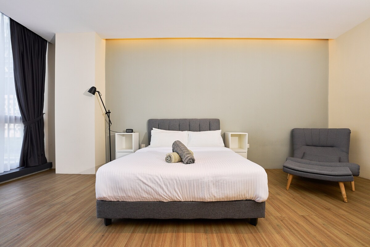 A spacious bedroom features a large bed with a neutral-colored headboard and crisp white linens. A comfortable armchair sits nearby, complemented by soft lighting against a muted wall. Light streams in through the floor-to-ceiling window, dressed with dark curtains.