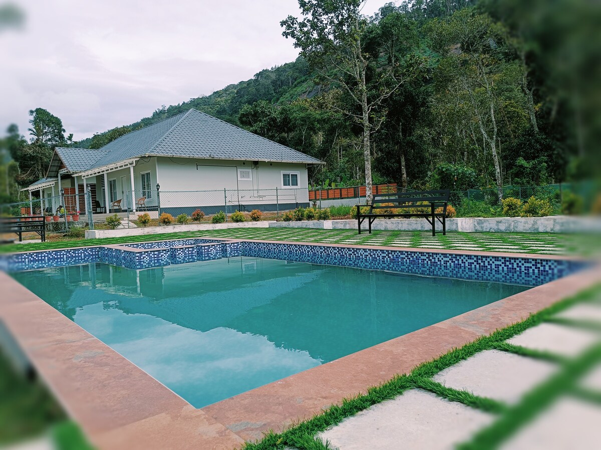 A clear pool reflects the surrounding greenery, featuring a mosaic-tiled edge. Nearby, a spacious house with a gabled roof is visible, set against a backdrop of lush hills. Benches offer seating around the pool area, enhancing the outdoor space.