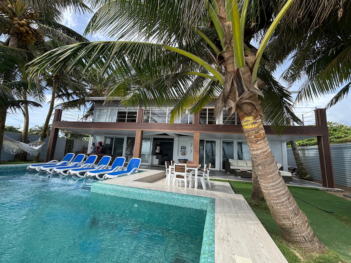 A spacious outdoor area features a shimmering pool surrounded by lounge chairs set under palm trees. A dining table is positioned nearby, with a hammock visible in the background. The house offers a light-colored exterior and large windows, providing a refreshing view.