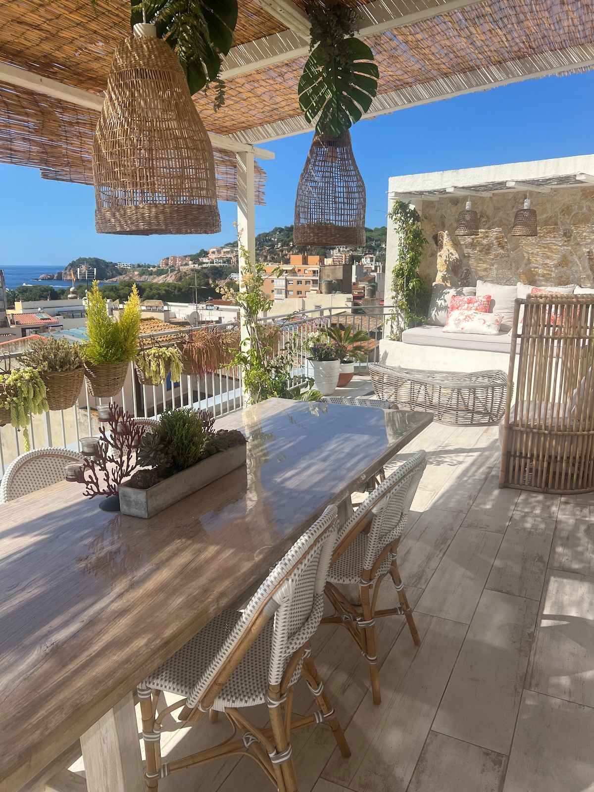 A spacious terrace is featured with a dining table surrounded by six chairs and adorned with potted plants. Stylish hanging lamps provide ambient lighting, while a lounge area with cushions invites relaxation. A view of the sea and cityscape is visible in the background.