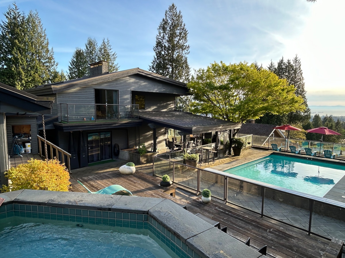 An expansive outdoor area showcases a large pool and hot tub, surrounded by a textured stone patio. Sun loungers and a dining area are positioned nearby, with shaded seating under an umbrella. The home features large windows, providing a view of the surrounding greenery.