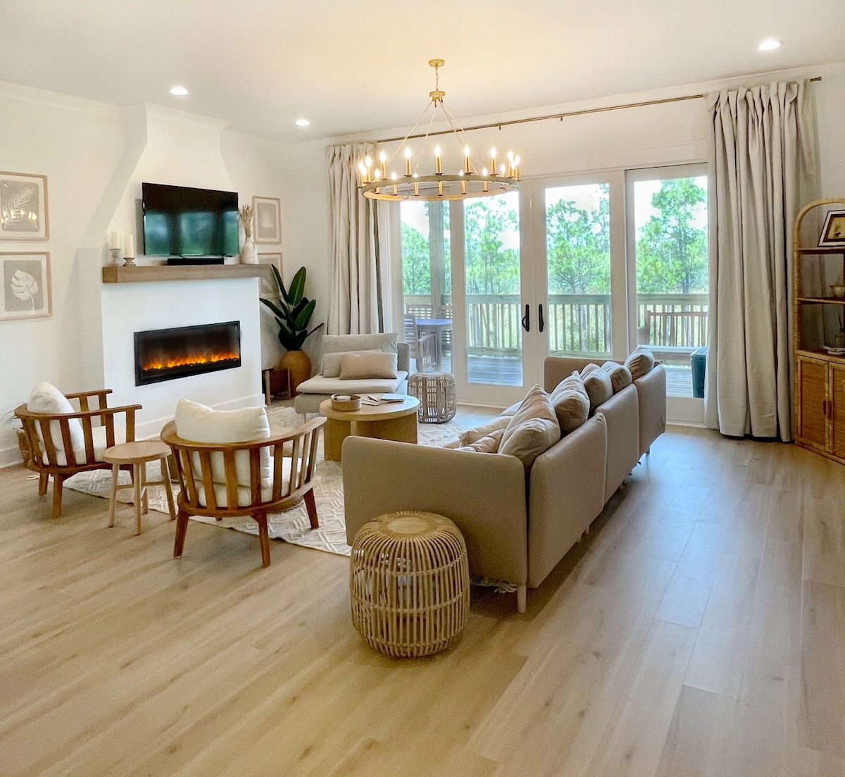The living area features an open layout with large windows that allow natural light to fill the space. A comfortable seating arrangement includes a gray sofa and two wooden chairs. A smart TV is mounted above a modern electric fireplace, surrounded by decorative elements and plants.
