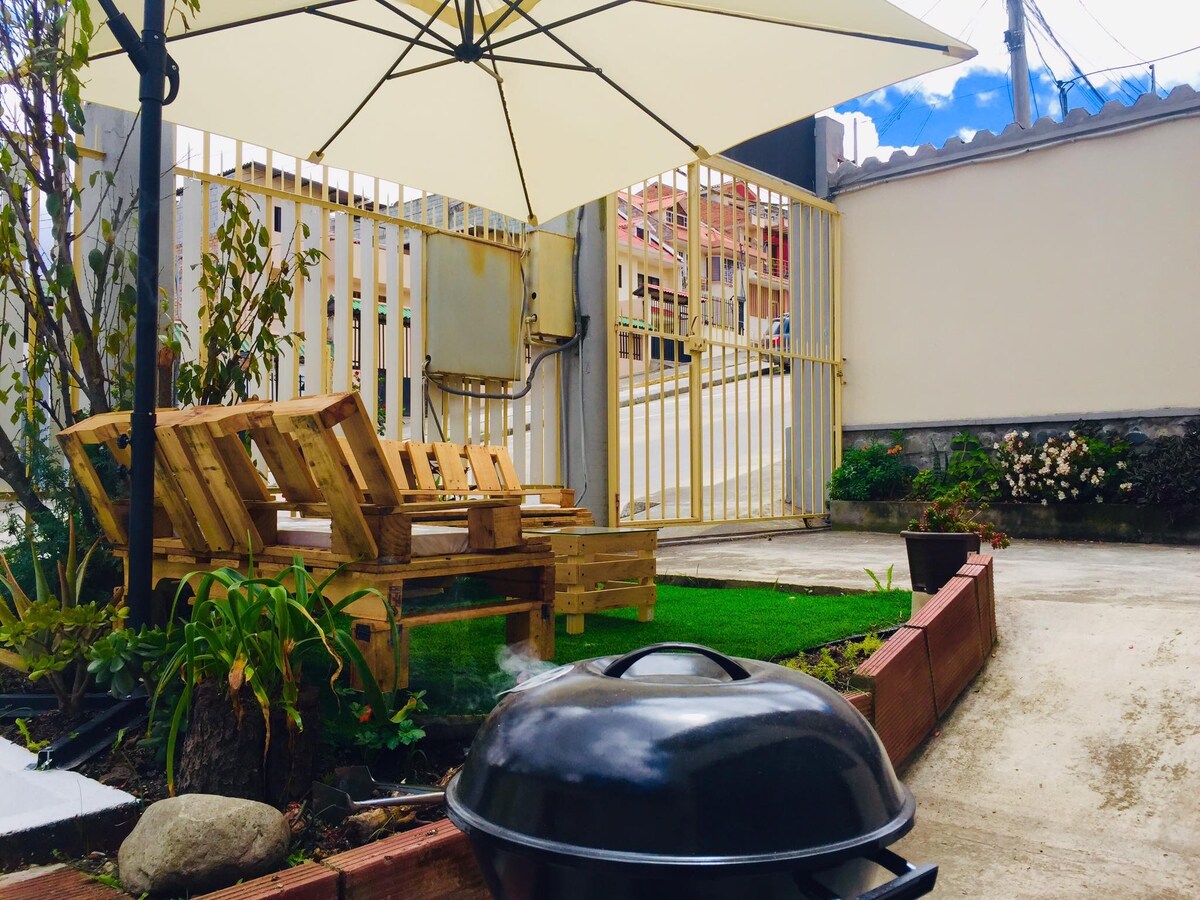 A small outdoor area features wooden lounge chairs positioned under a large umbrella. A charcoal grill is situated on a stone pathway, surrounded by a small garden with green grass and plants. A gated entrance provides access to the peaceful space.