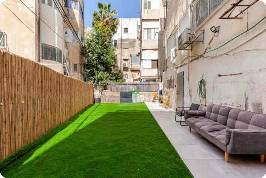 A spacious outdoor garden area is visible, featuring well-maintained artificial grass. A comfortable gray sofa is positioned against a wall, providing a relaxed seating option. Surrounding buildings contribute to a private ambiance, with tall trees adding greenery to the setting.