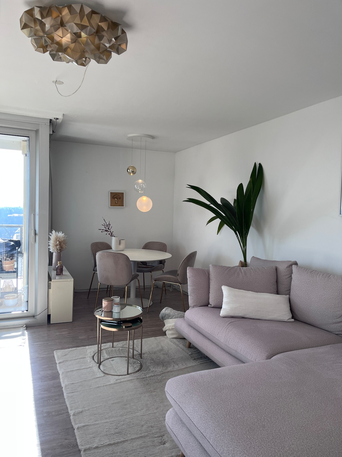 A bright living room is presented with a spacious sofa and elegant accent chair. A small dining table with four chairs is positioned near a glass sliding door. Natural light fills the space, highlighting a potted plant and tasteful decorative elements.