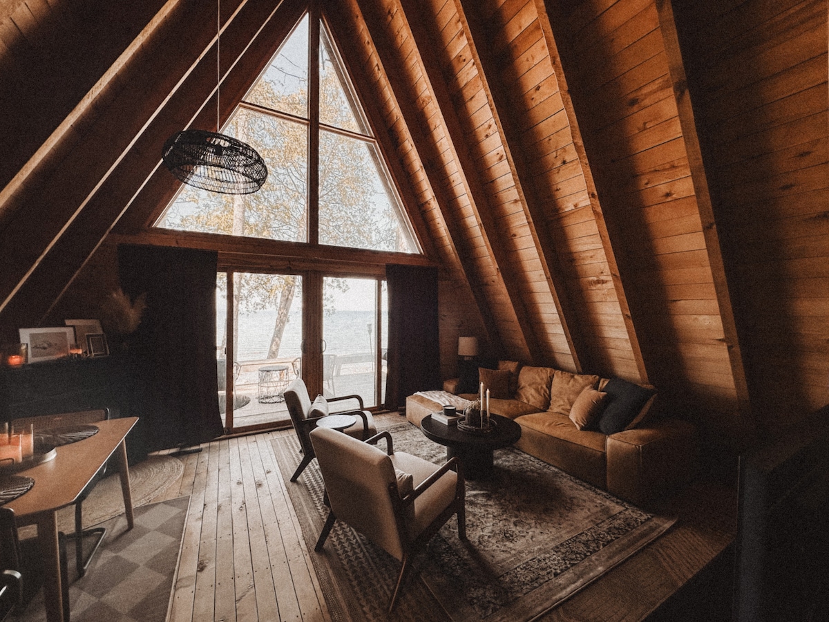 An A-frame cabin living room features tall wooden walls and a vaulted ceiling. Large windows provide views of Lake Huron, allowing natural light to fill the space. A comfortable sofa, two armchairs, and a round coffee table are arranged thoughtfully. A dining area is visible in the background.